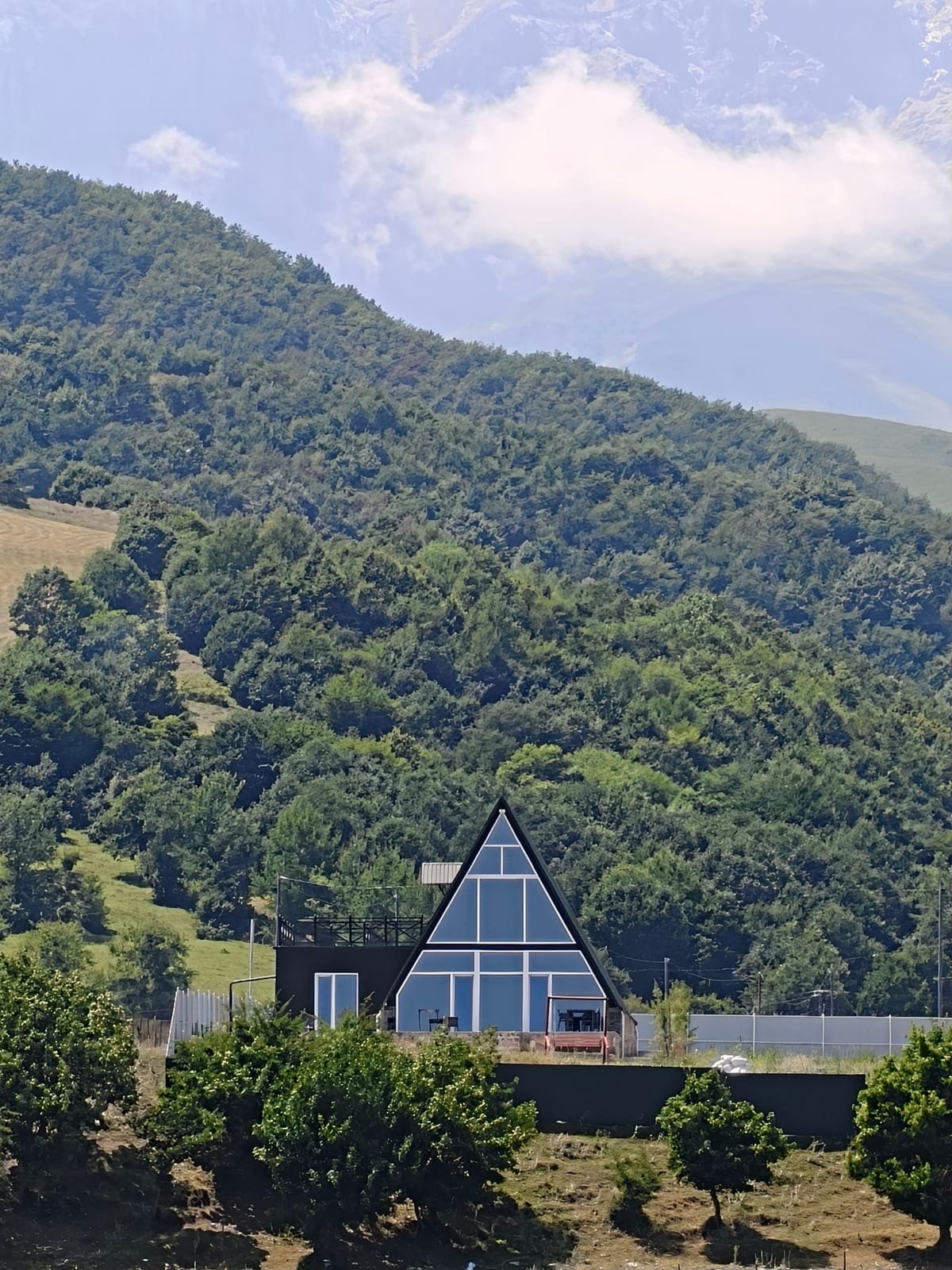 The triangular SkyHouse seen from a distance against a wooded slope and mountains in cloud