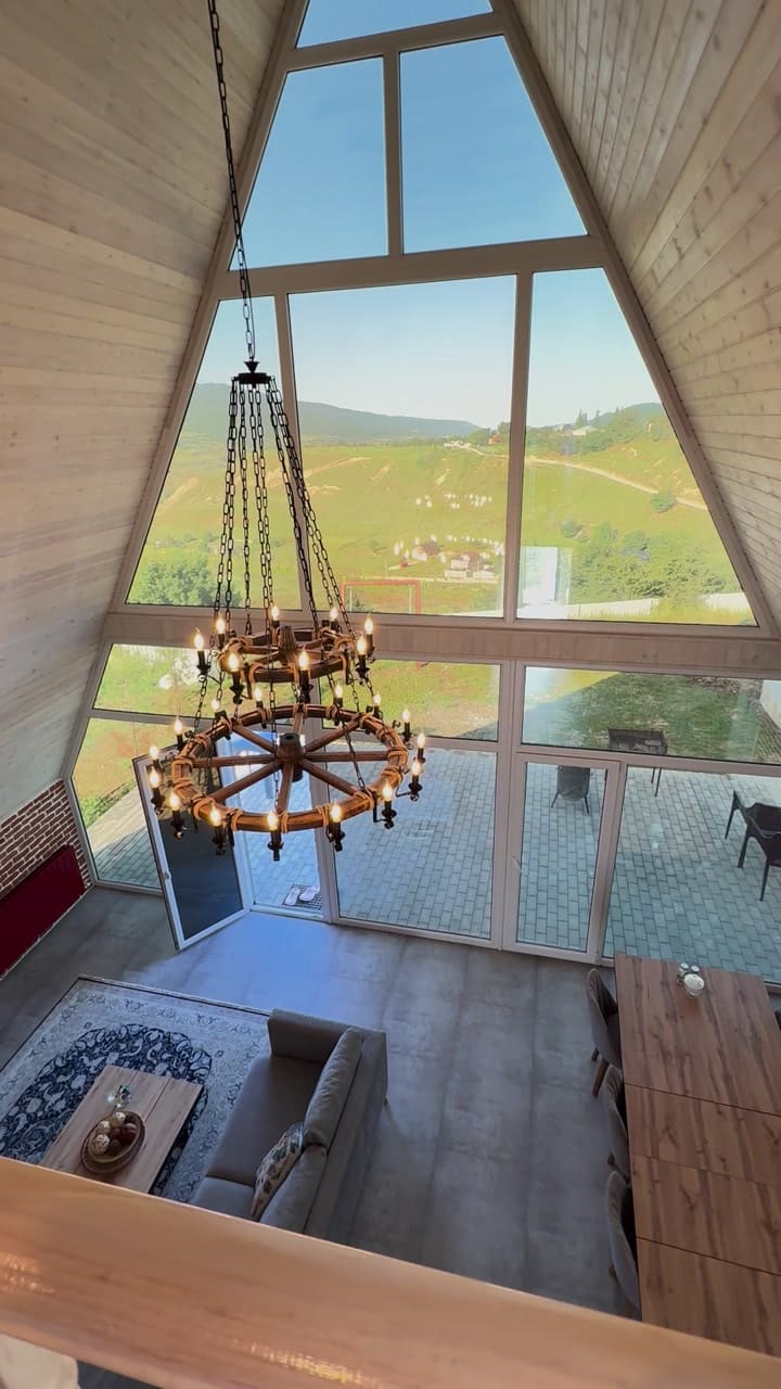 View from the upper level into the living room: a triangular panoramic window, a chandelier and mountains beyond the glass